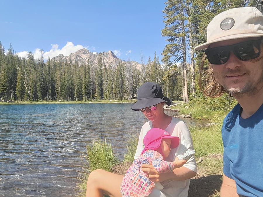 a family of 3 pose for a photo at the top of the alpine lake hike in the sawtooth mountiains. the lake and alpine scenery is in the background.