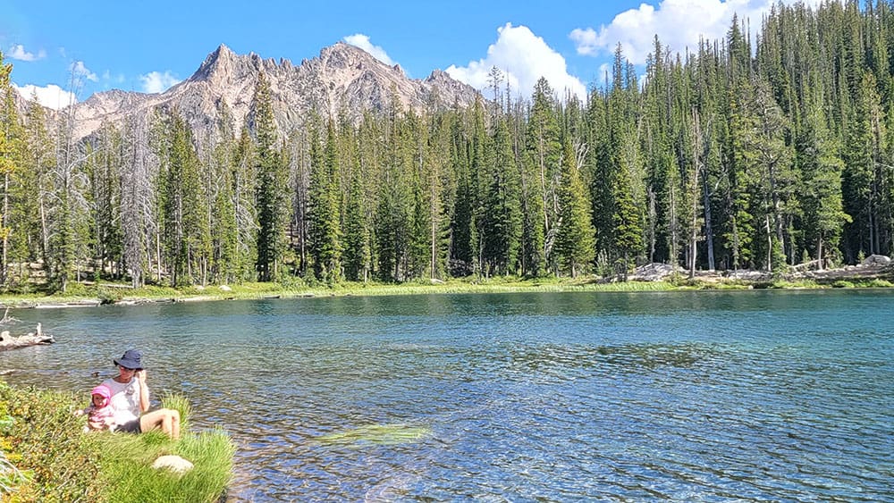 a woman and her baby sitting beside an alpine lake in the sawtooth mountains of idaho