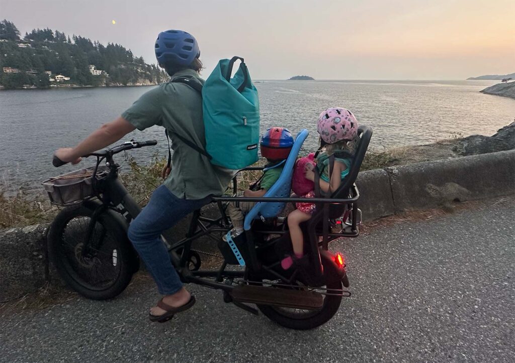 a father and two children look out at the sunset from the edge of whytecliff park while seated on a cargo e-bike
