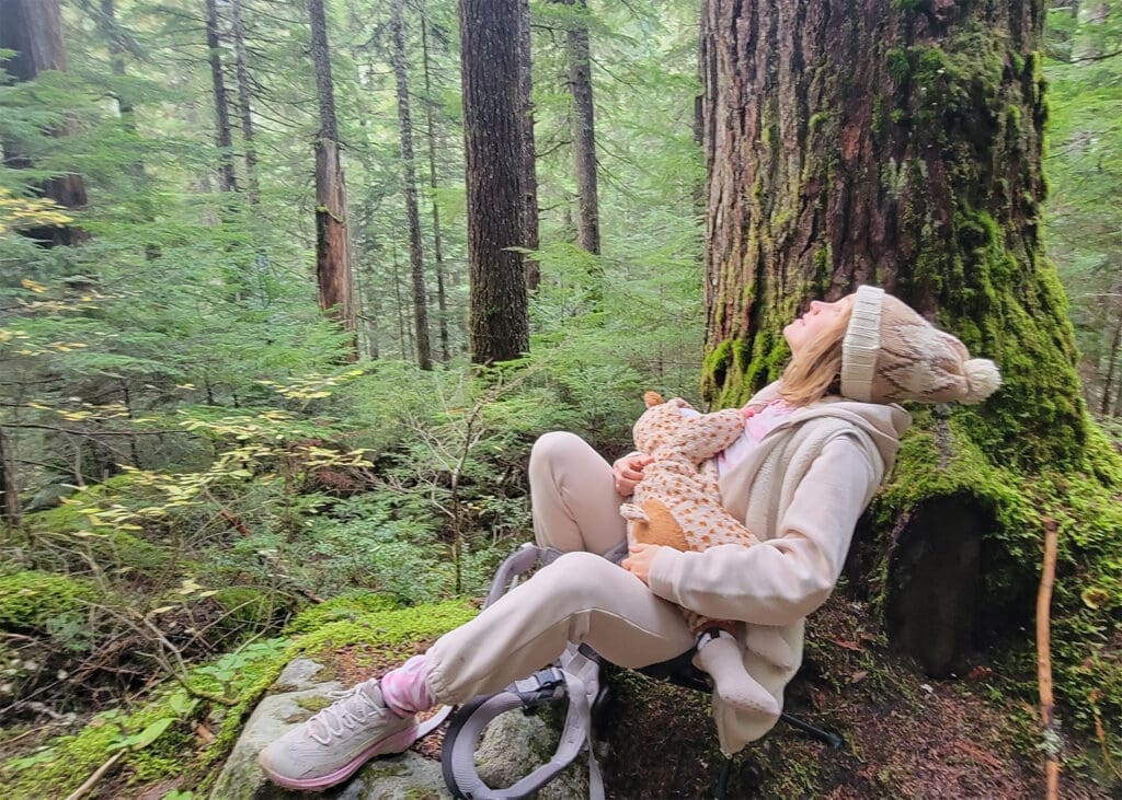 a woman breastfeeds her baby while next to a large douglas fir tree on the trail to cheakamus lake in garibaldi provincial park.