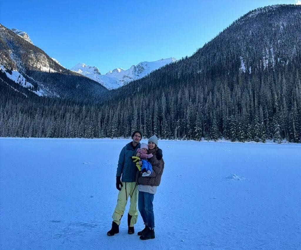 a mother, father, and baby pose for a photo while standing on the lake at Joffre Lakes Park in the winter