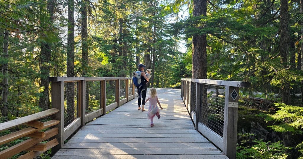 a young girl runs on a bridge while her mother carries her baby brother while walking ahead in the forest