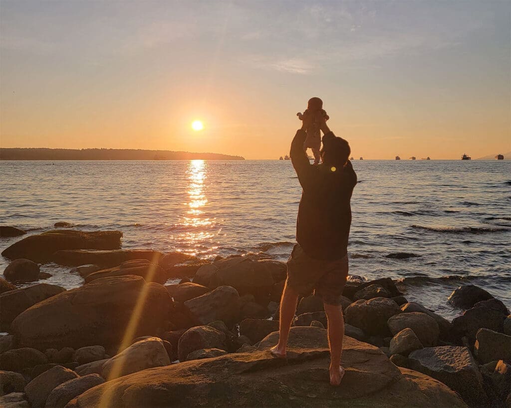 a father raises his baby into the sky at second beach in vancouver, looking at the sunset